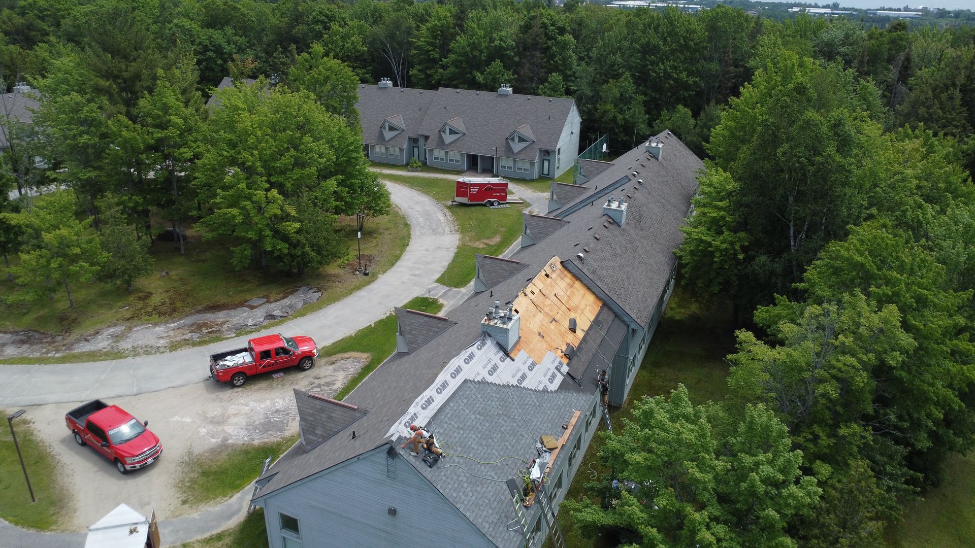 Aerial view of gray houses along a winding driveway, with two red vehicles and dense green trees.