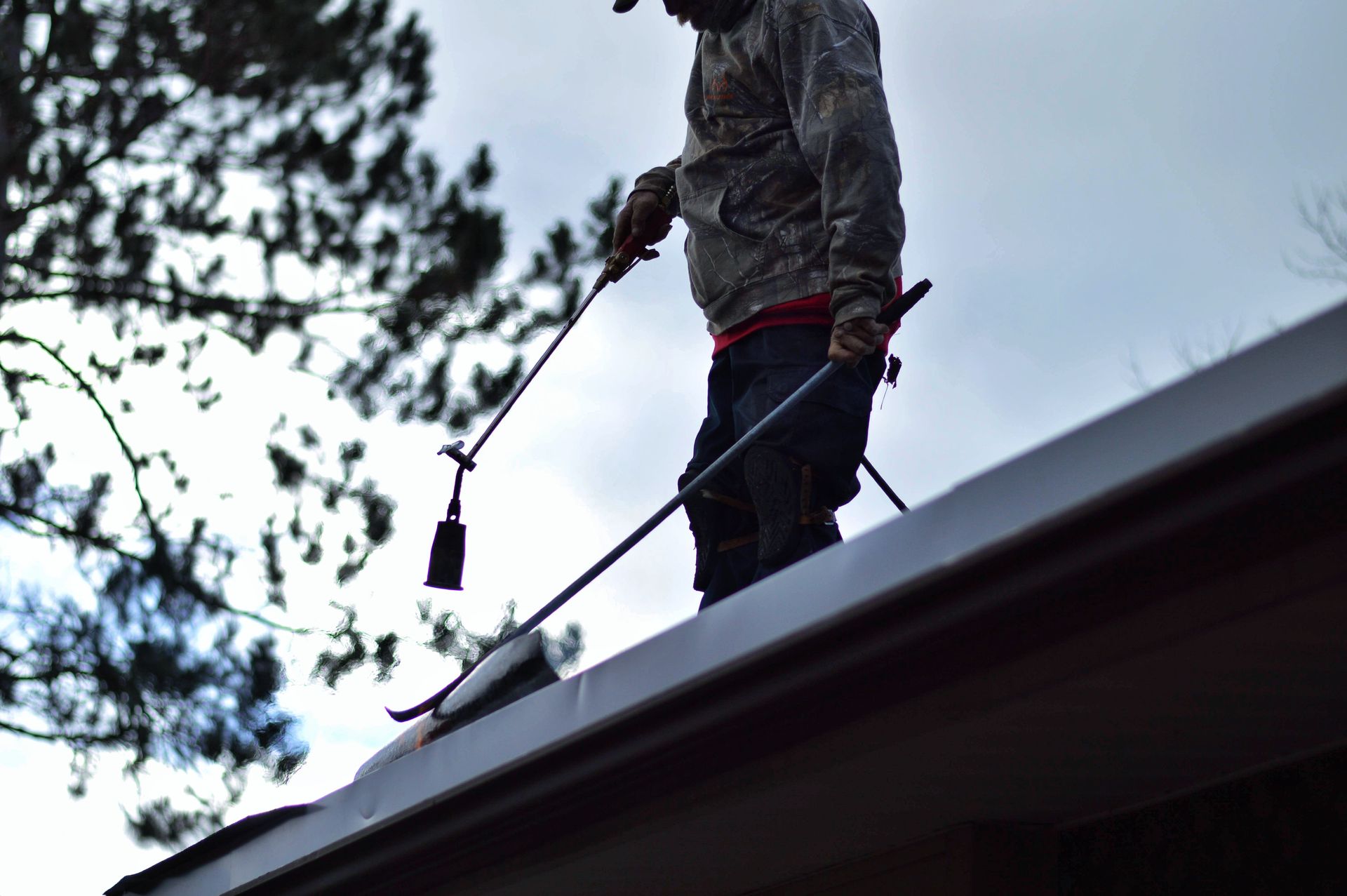 Person on roof cleaning gutter with long-handled tool, cloudy sky, trees in background.