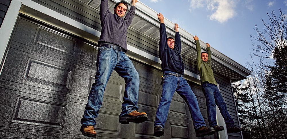 Three people hanging from a garage door's rain gutter against a blue sky.
