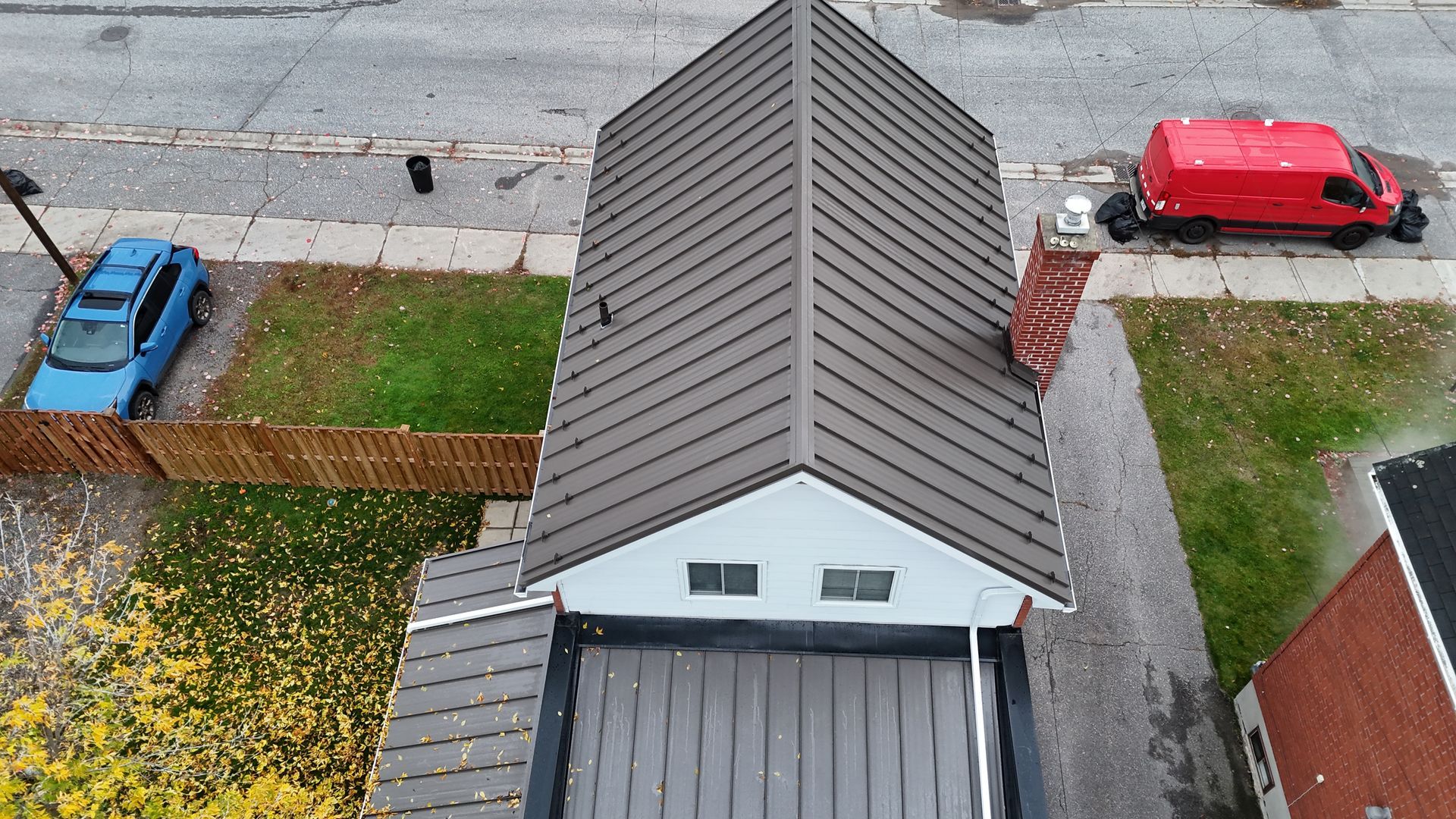 Aerial view of a house roof with a red car and blue car parked by the street, bordered by lawns and a wooden fence