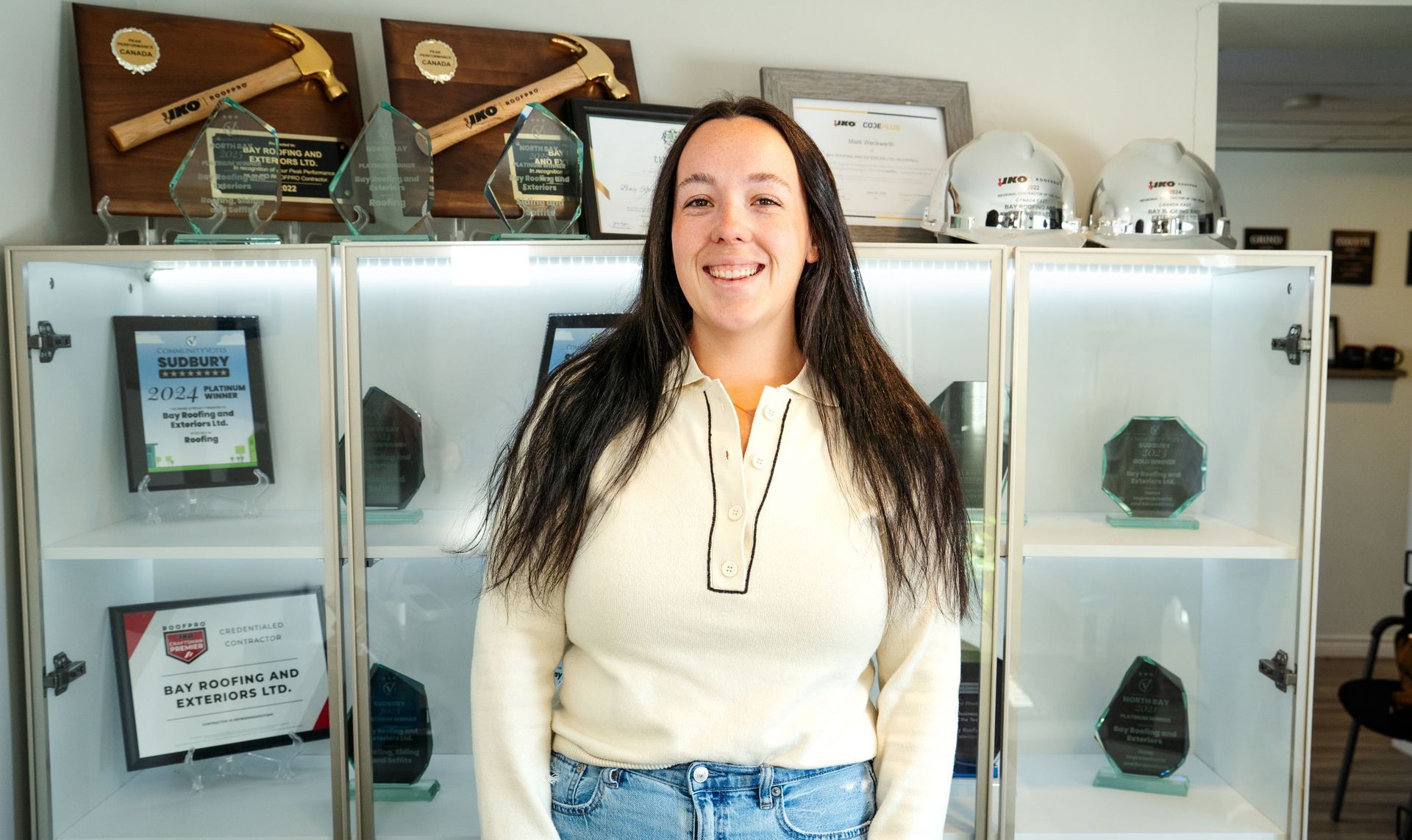 Woman in cream shirt smiling, standing in front of glass display case with awards and hard hats.