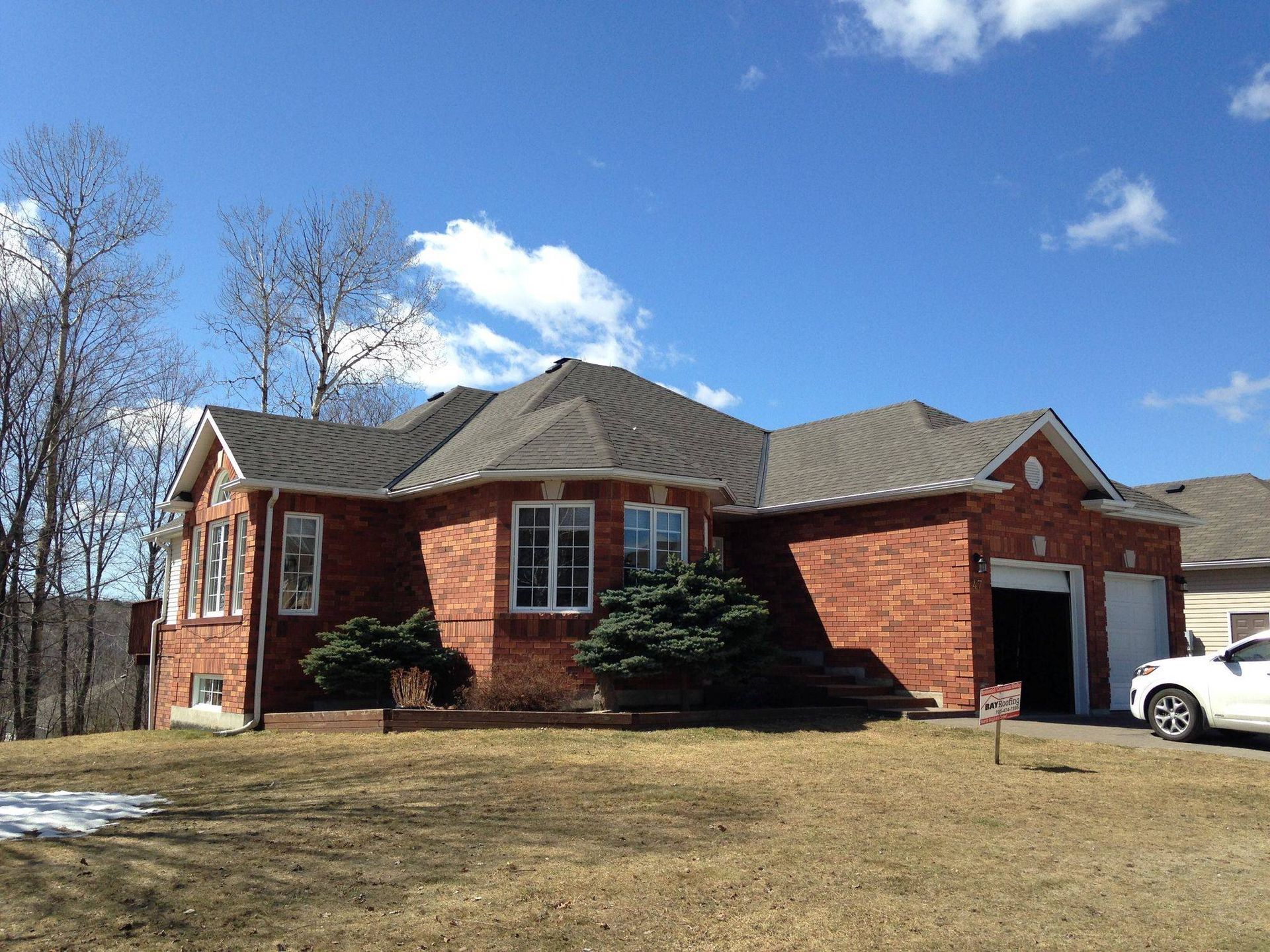 Brick house with a gray roof and white garage under a blue sky, with a car parked in front.