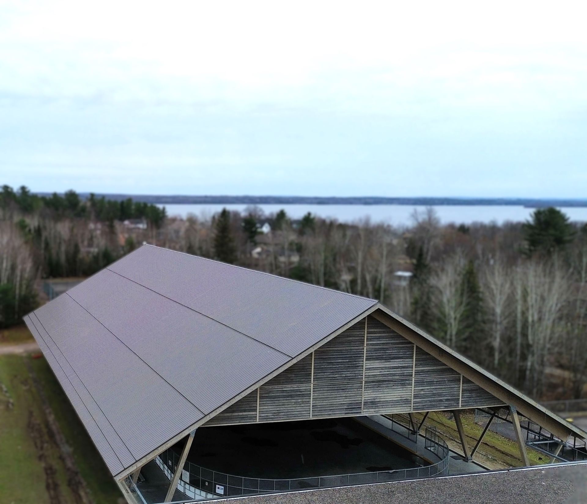 Aerial view of a large wooden barn with a gray roof beside forest and a lake