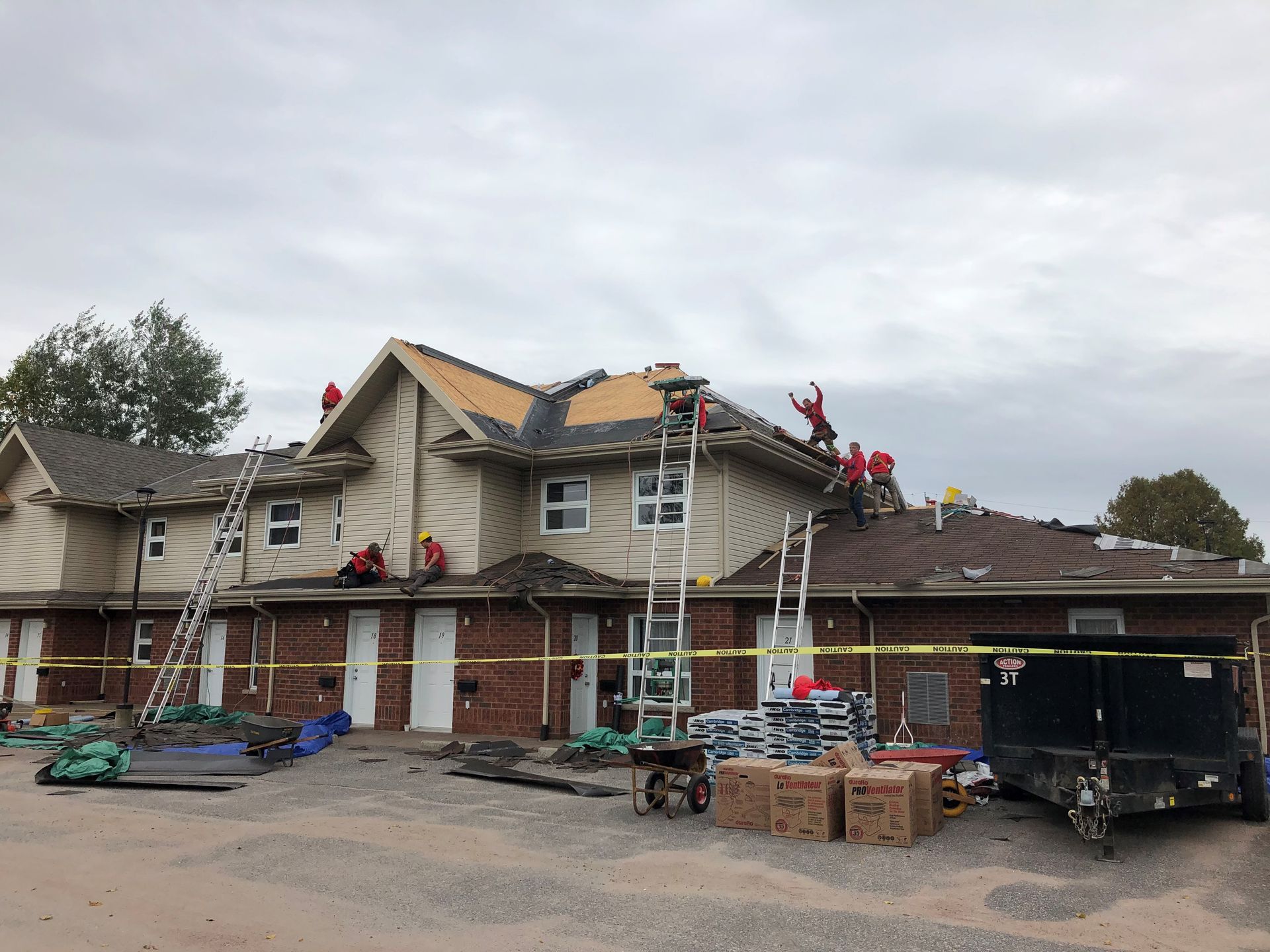 House roof under repair with ladders, workers, and construction equipment in front of a two-story building