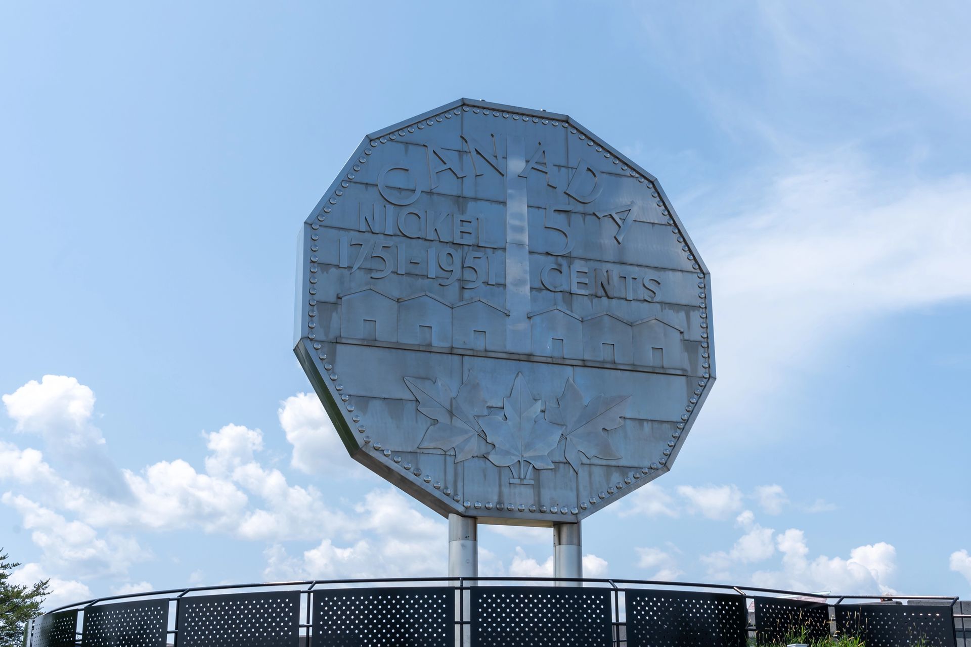 Large octagonal roadside sign against a blue sky with clouds, mounted on white posts above a fence