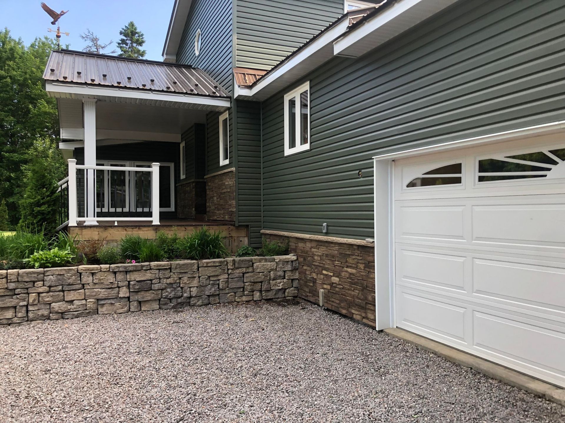 Gravel driveway beside a green house with white garage door and stone retaining wall