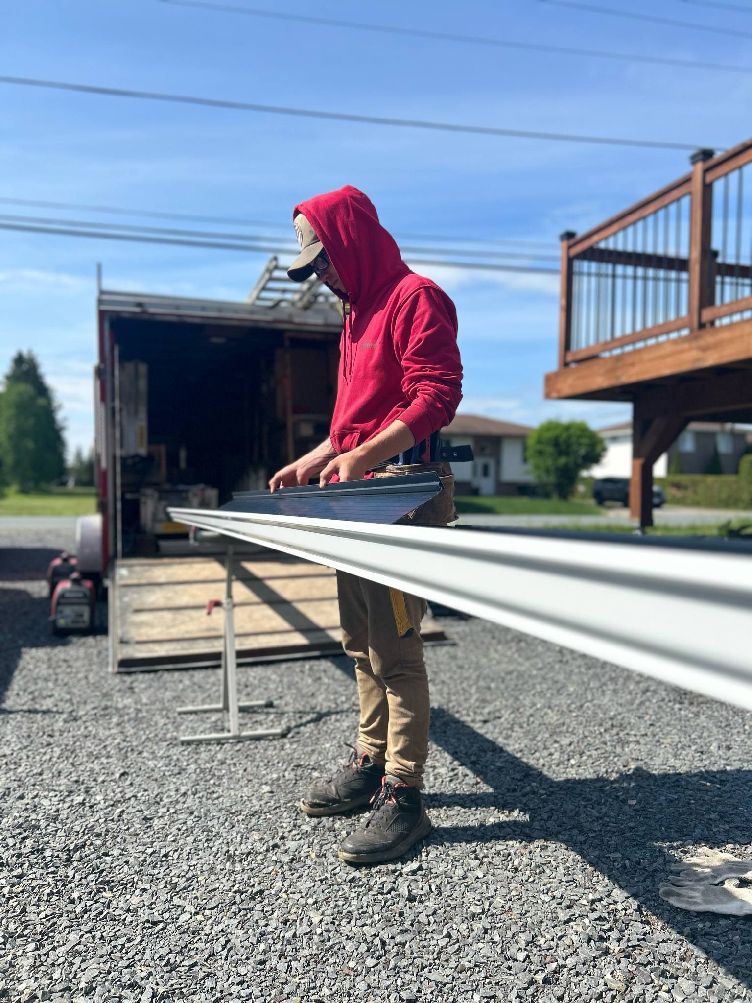 Worker in red hoodie carrying a long metal beam beside a trailer on a gravel driveway.