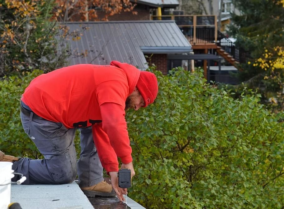 Roofer in red hoodie kneeling, working on a flat roof. Brown boots, gray pants, green trees in background.