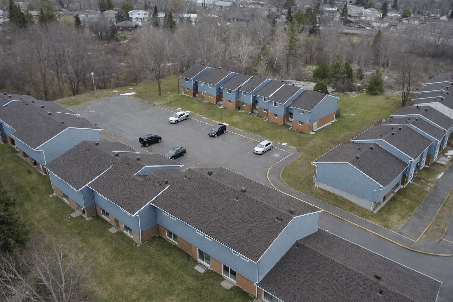 Aerial view of a blue-gray apartment complex with parked cars and trees surrounding the buildings