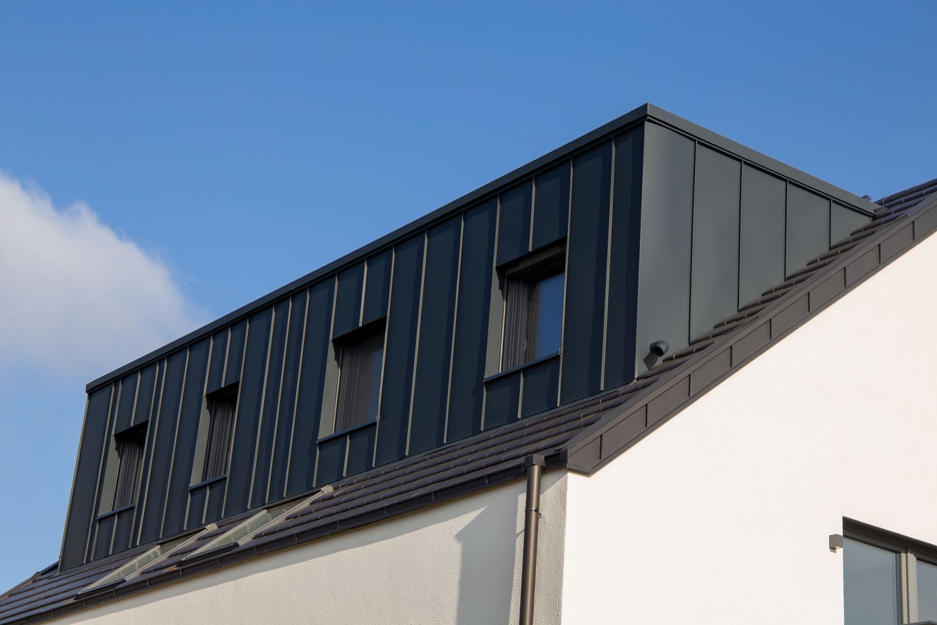 A modern house with dark gray metal roofing and dormer windows against a blue sky.