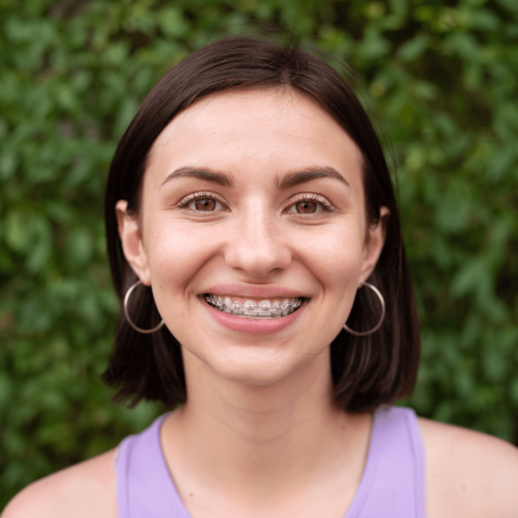 Smiling person with braces in a purple top, standing outdoors against green foliage