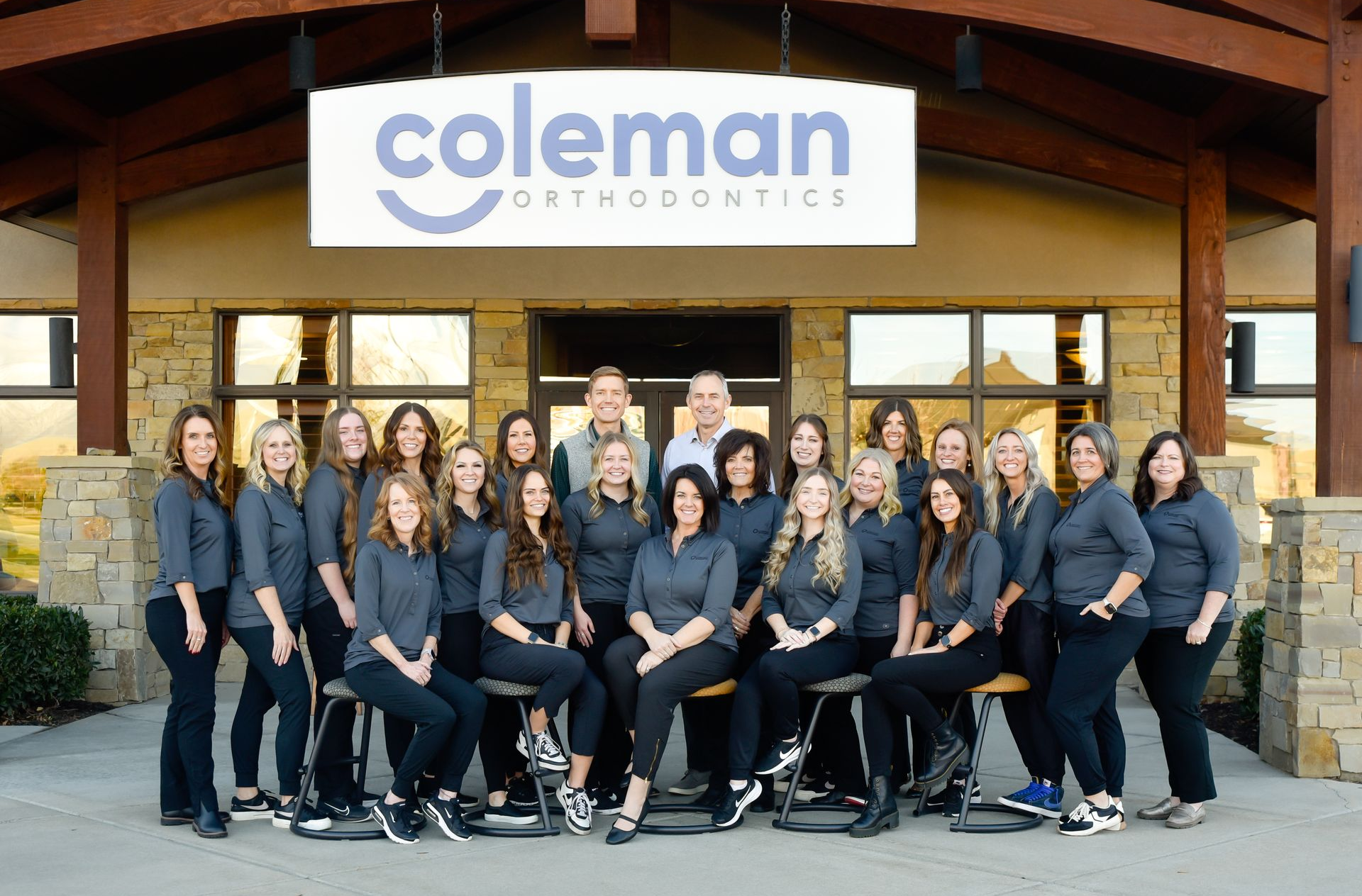 Group photo of Coleman Orthodontics staff in front of their office. They are wearing gray shirts and black pants.