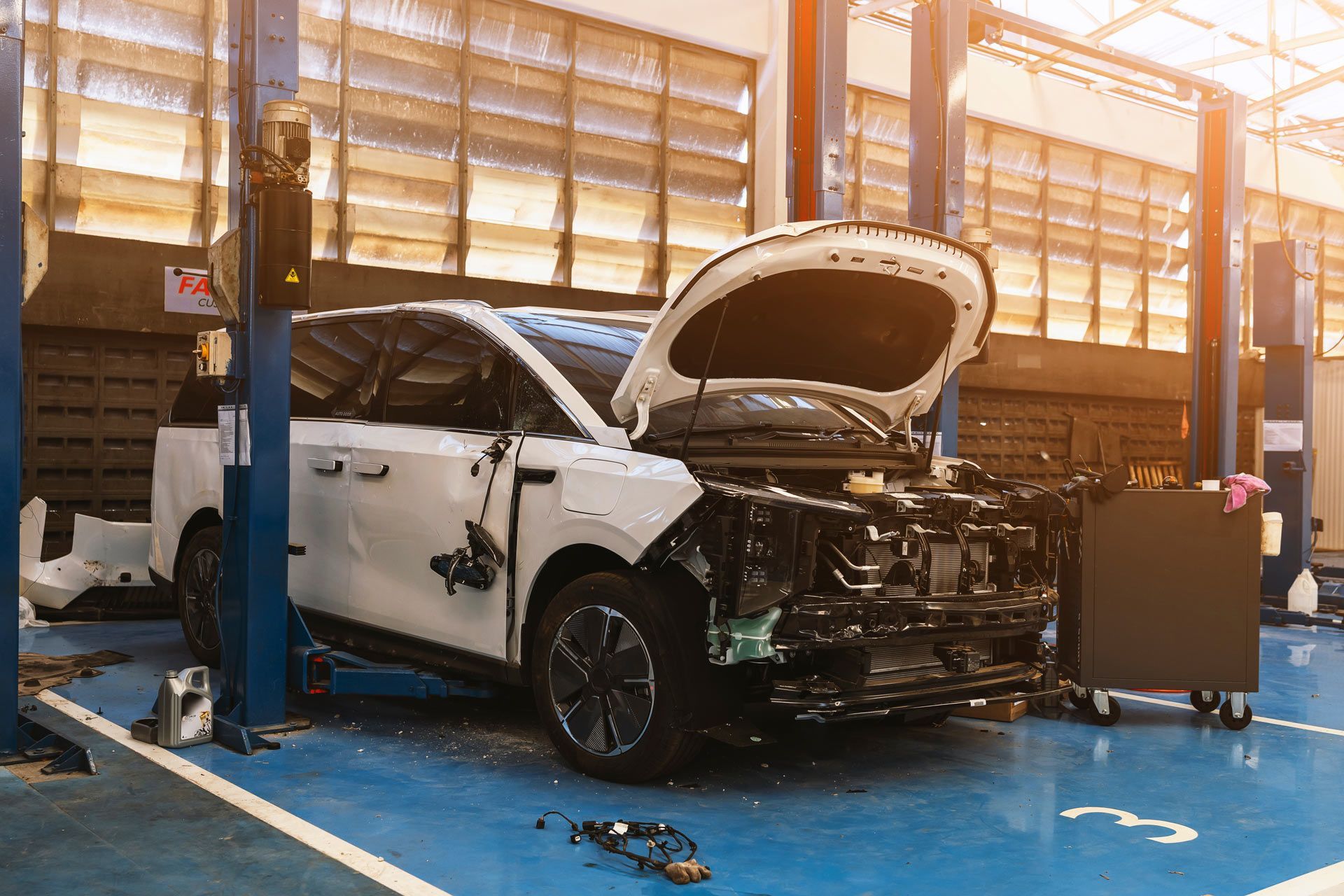 A white minivan with its hood and front bumper removed is undergoing repairs in an auto shop with blue floors.