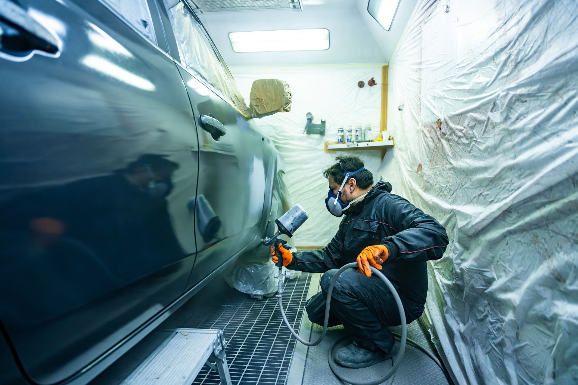 A person in a protective suit and respirator sprays paint on a car inside a well-lit auto body workshop.
