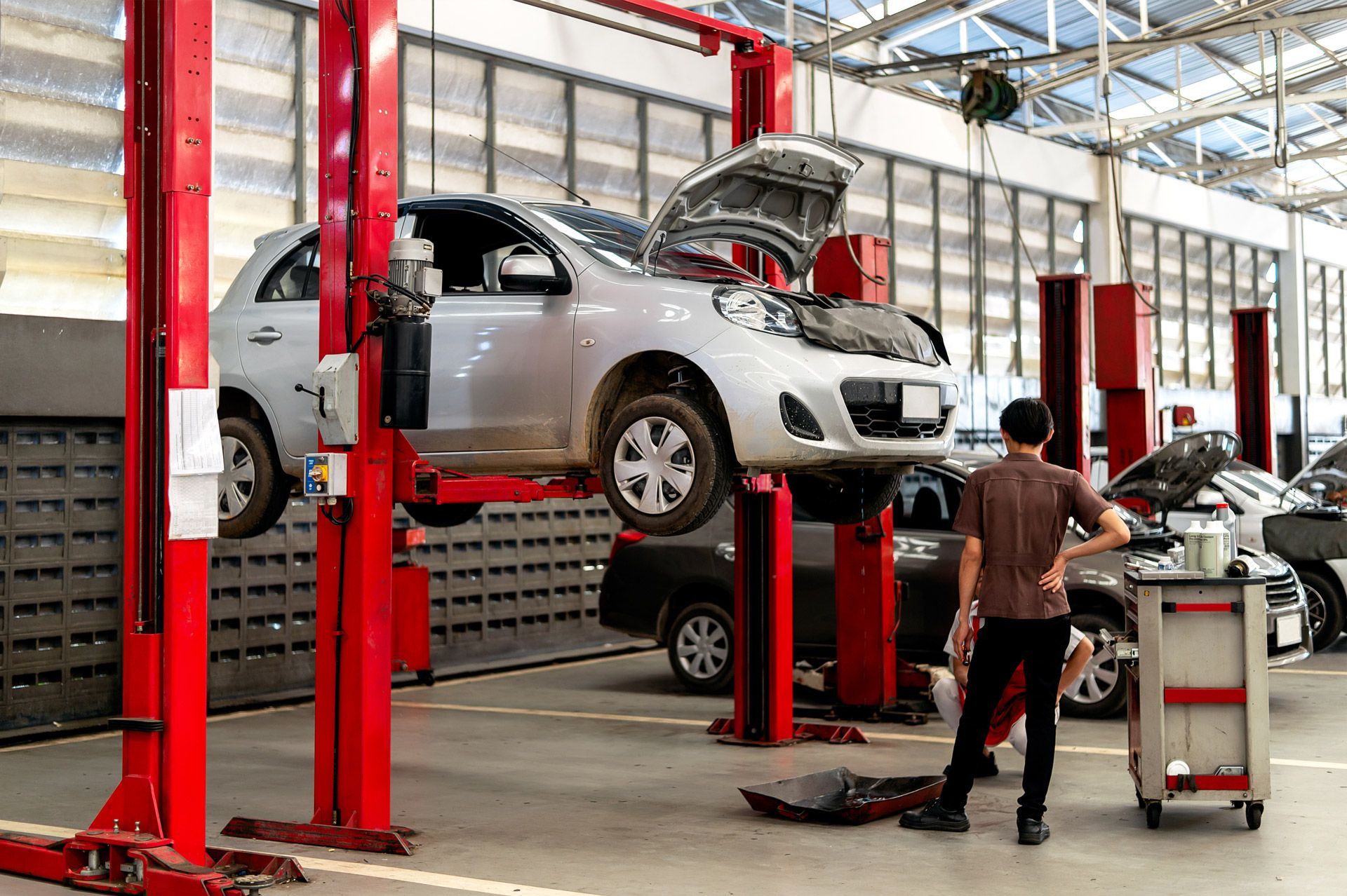 A silver car with its hood up is lifted on a red hydraulic hoist in an auto repair shop while a mechanic stands nearby.