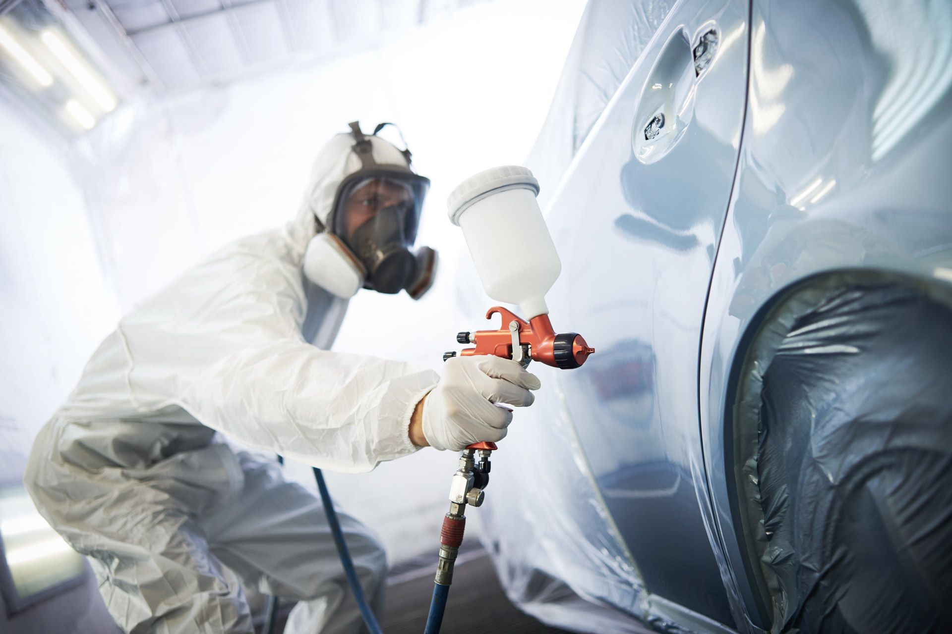 A person in a protective suit and respirator sprays paint onto a light blue car inside a workshop.
