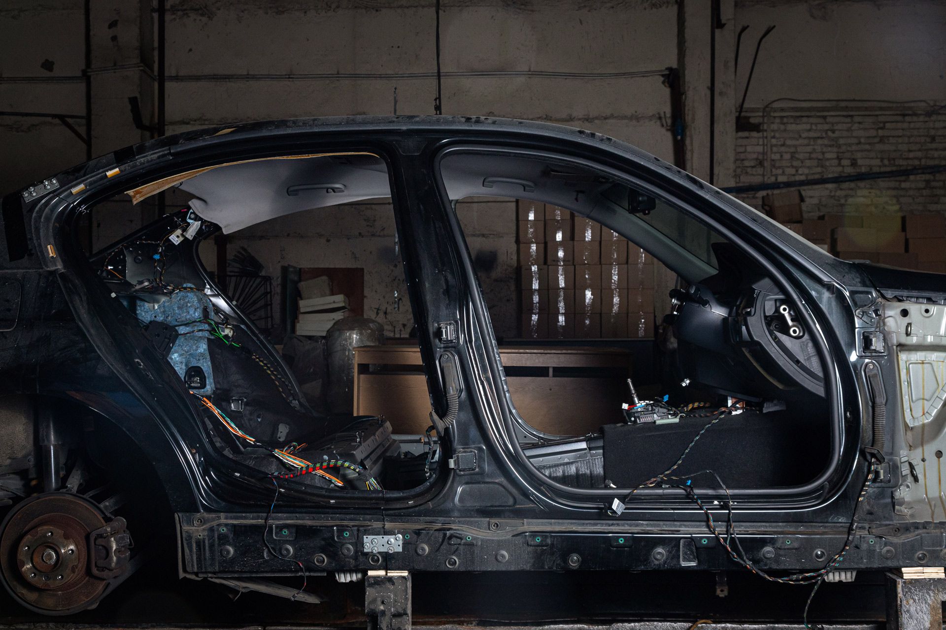 A disassembled black car frame sits on wooden blocks inside a dimly lit workshop, missing doors and interior parts.