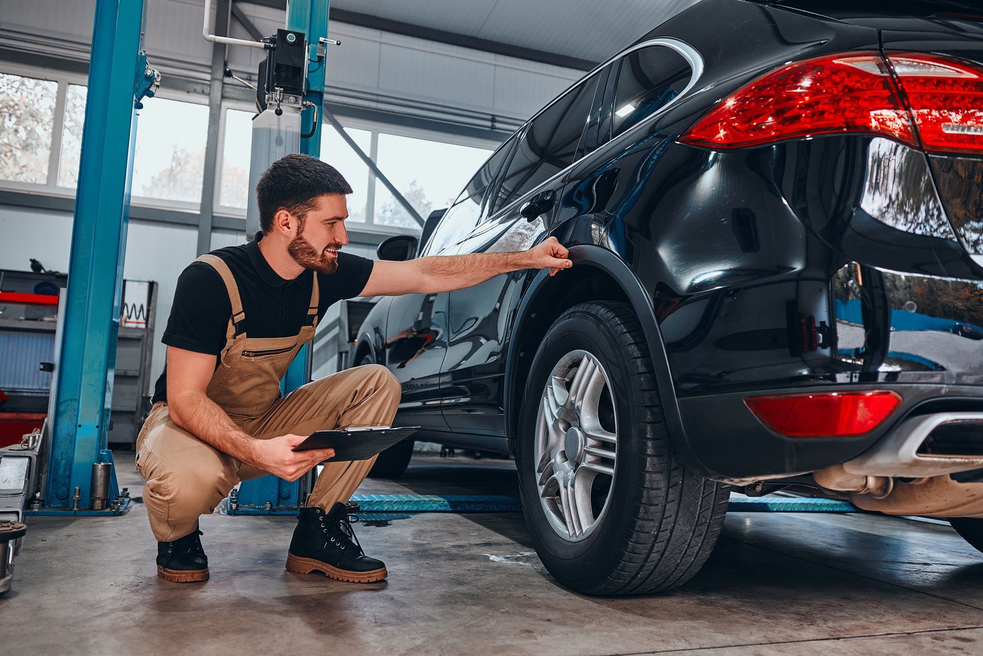 A mechanic in tan coveralls crouches by a black car in a garage, inspecting the wheel well while holding a clipboard.