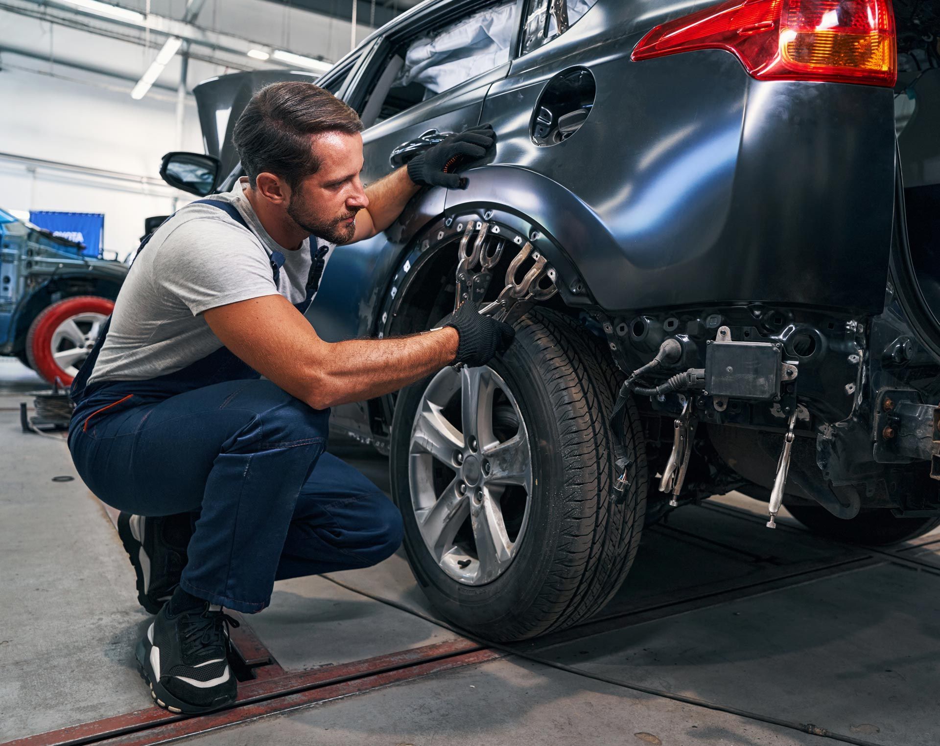 A person in a white t-shirt and dark overalls kneels to repair the rear wheel arch of a matte-black car in a workshop.