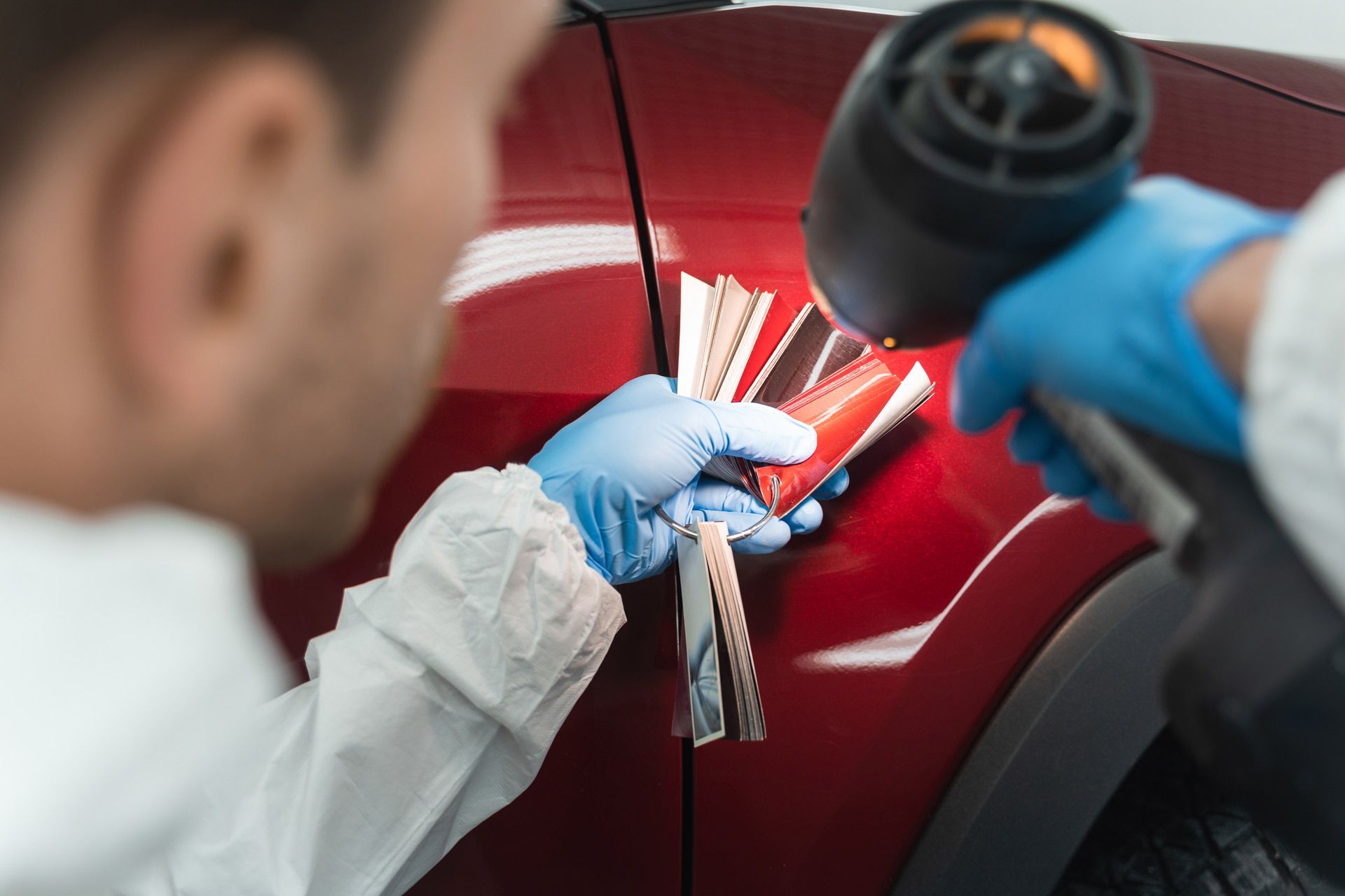 A worker wearing blue gloves uses a heat gun to match a car's red paint color using a fan deck of color samples.