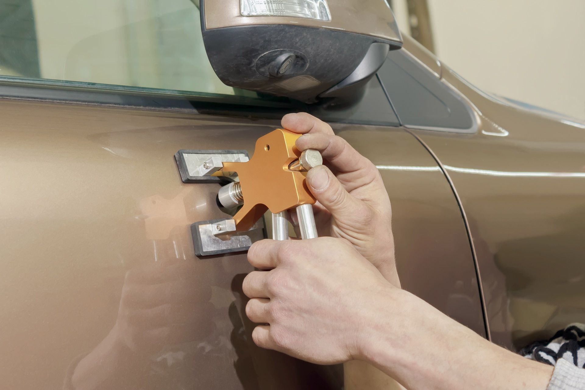 A hand uses a gold paintless dent repair tool on the brown door panel of a car.