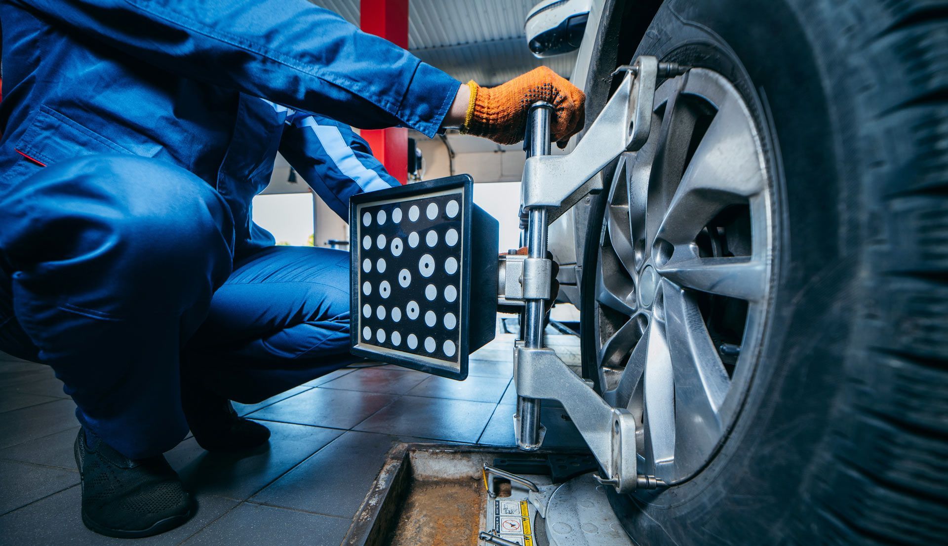 A mechanic in a blue uniform attaches a wheel alignment sensor to a vehicle's tire in a service garage.