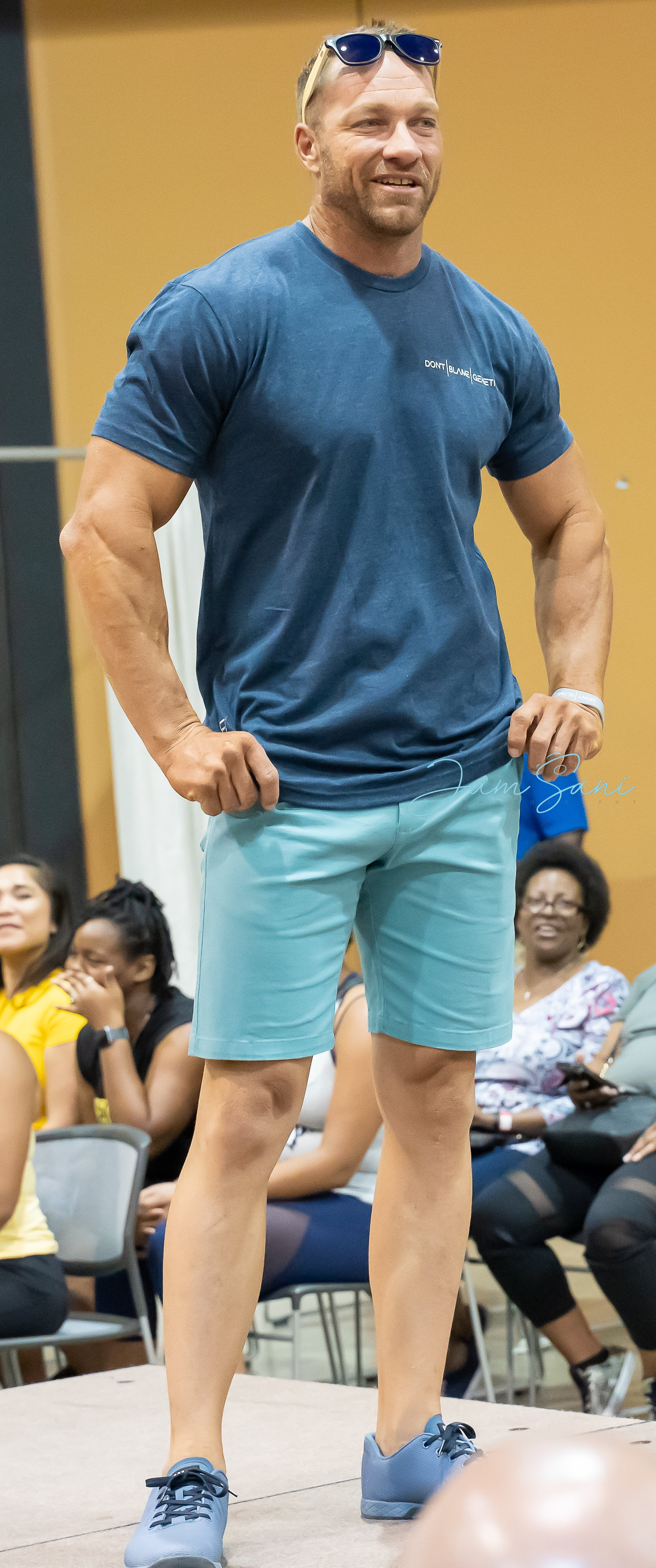 Man in blue T-shirt and teal shorts standing on a runway, with an audience seated behind him.