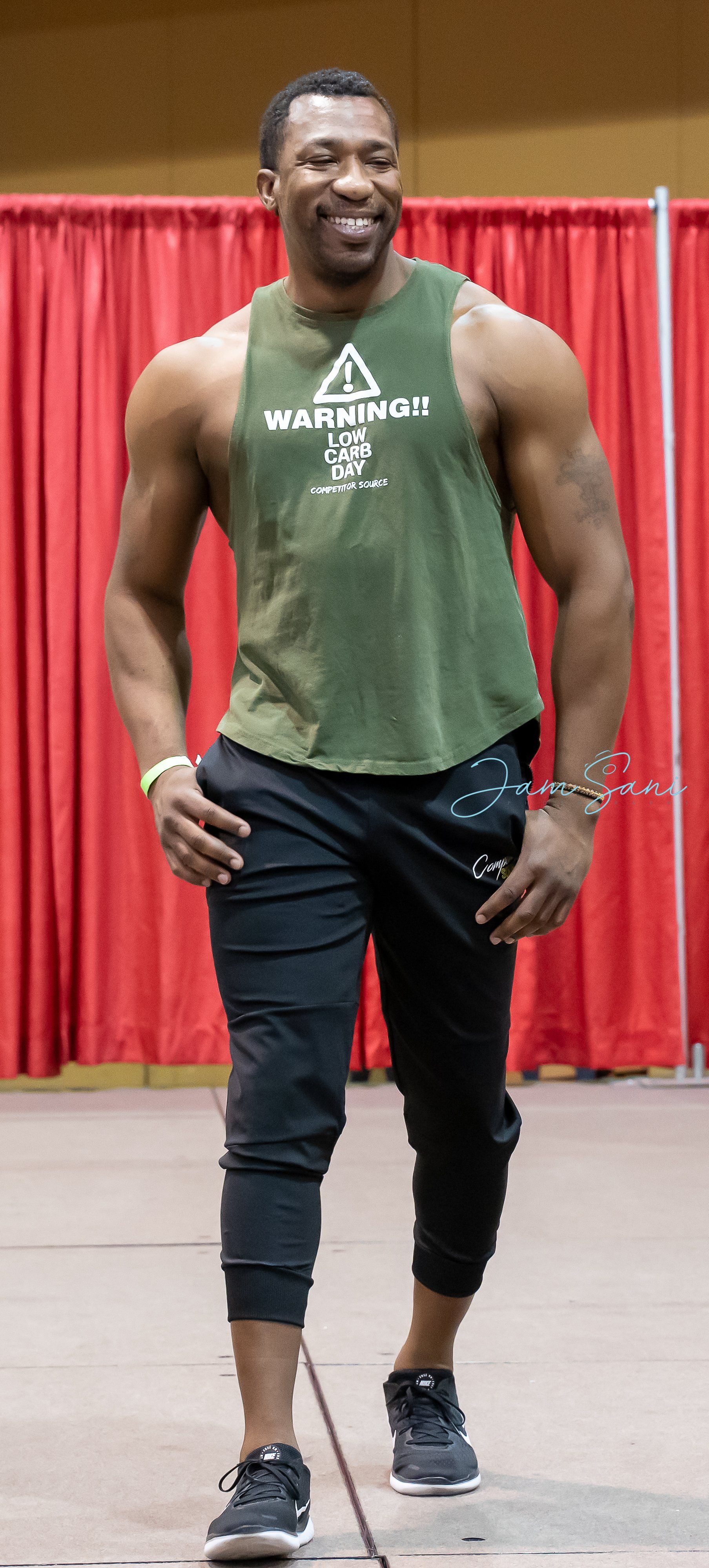 Man in green tank top and black joggers walking in front of red curtains on an indoor stage