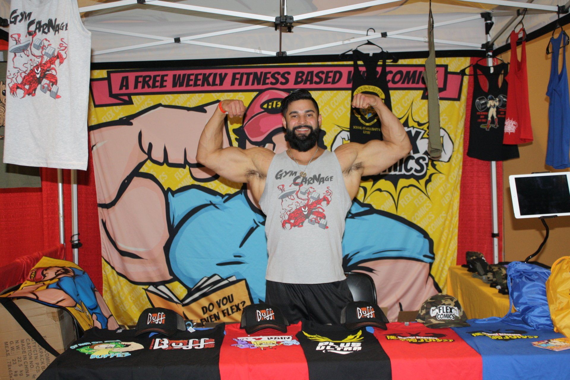 Man flexing behind a table at a fitness expo booth with colorful banners and shirts displayed