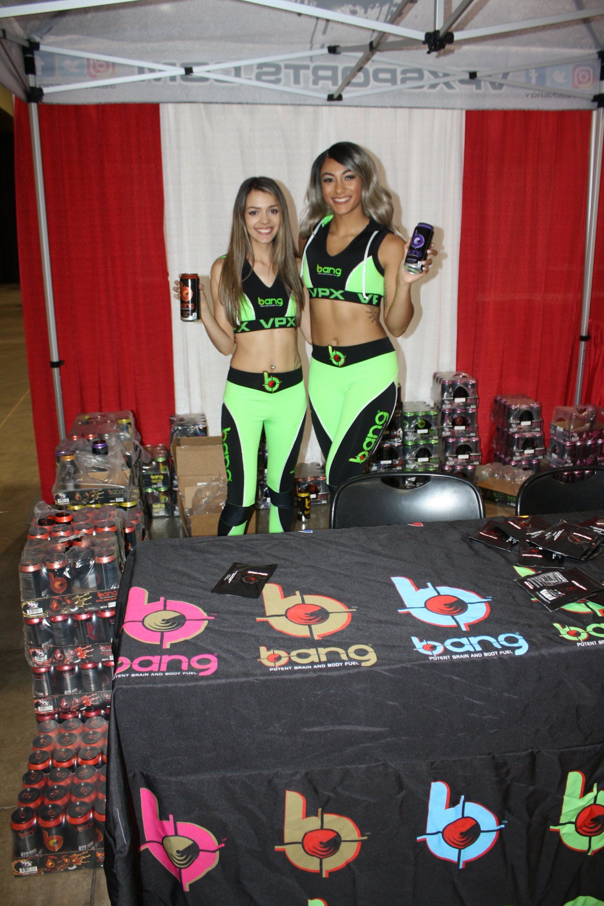 Two women in neon green outfits stand behind a branded table at an indoor event booth.
