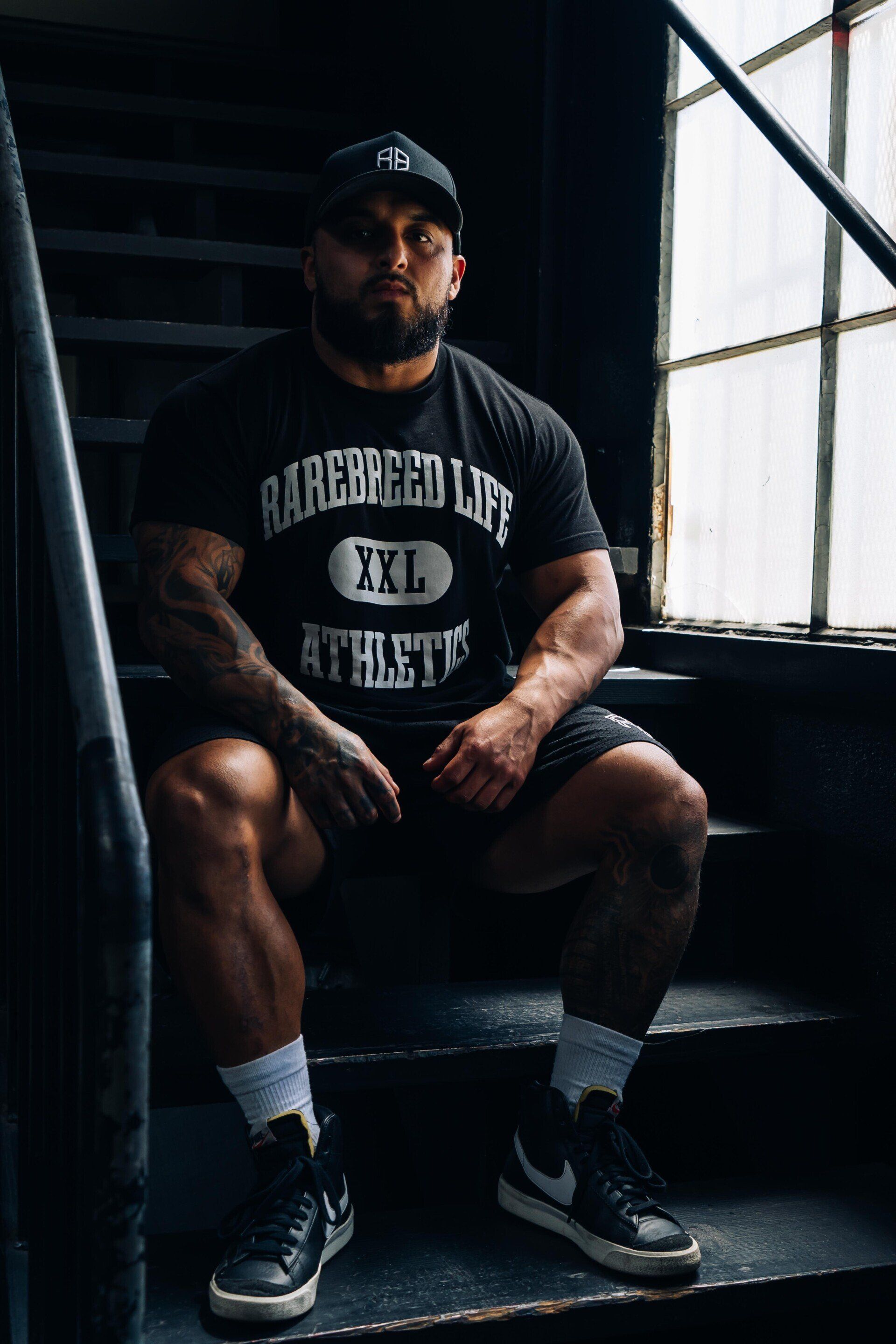 Person seated on stairs in a dim room, wearing a dark cap, black T-shirt, shorts, and sneakers.