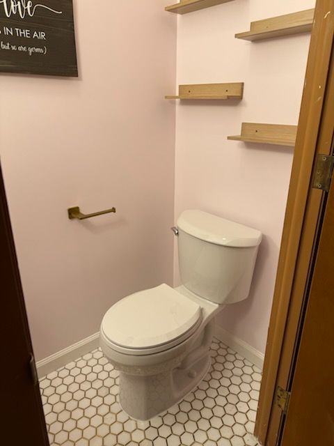 Pink-walled bathroom with white toilet, hexagonal tile floor, and wooden shelves. Brass toilet paper holder.