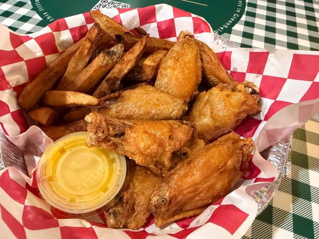 Chicken wings and fries in a basket with dipping sauce, red and white checkered liner, on a green table.
