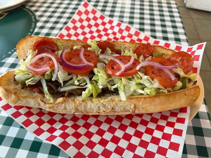Submarine sandwich on checkered paper, topped with lettuce, tomatoes, and red onions.