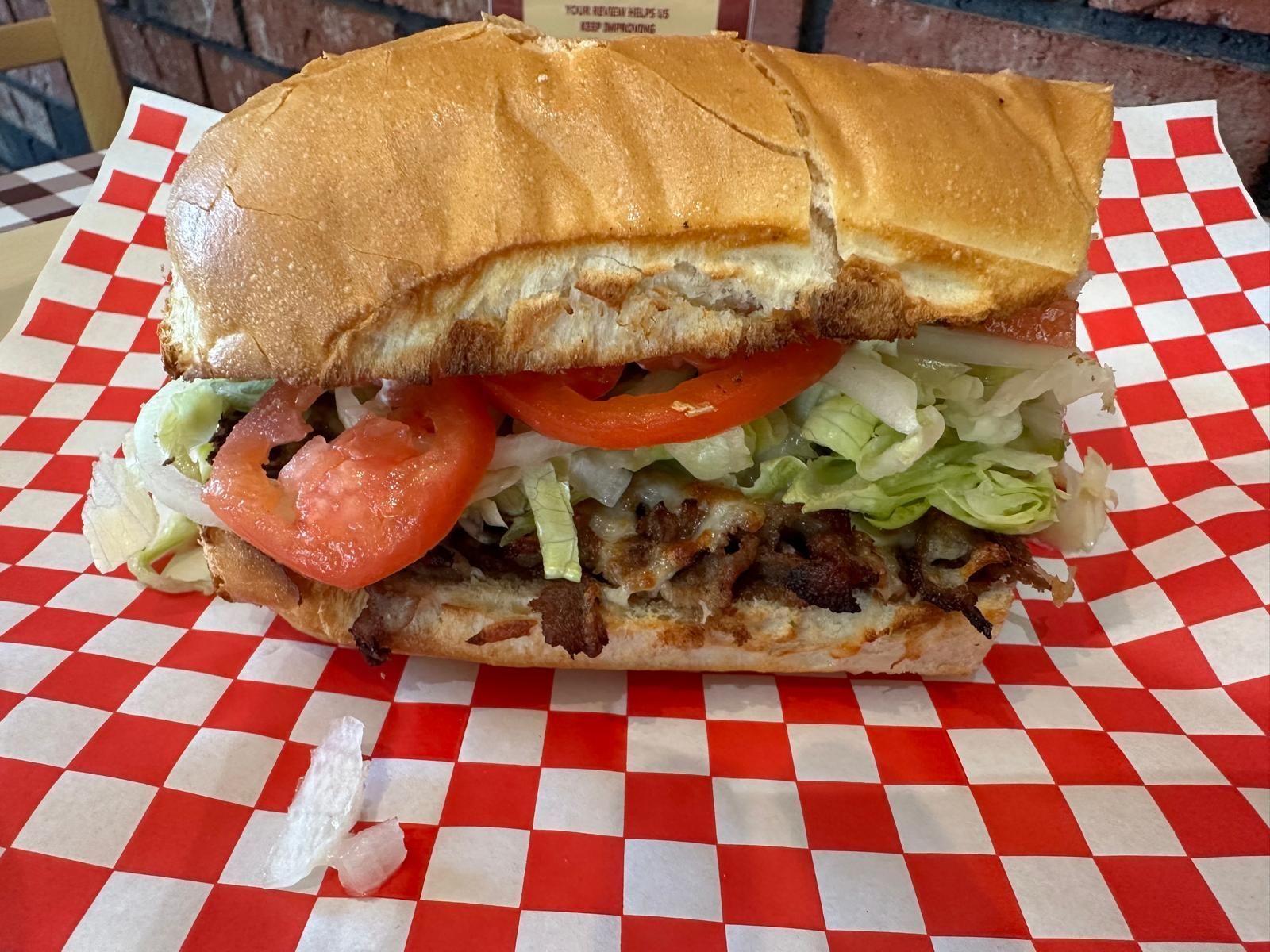 Sub sandwich with lettuce, tomato, and meat on red checkered paper.
