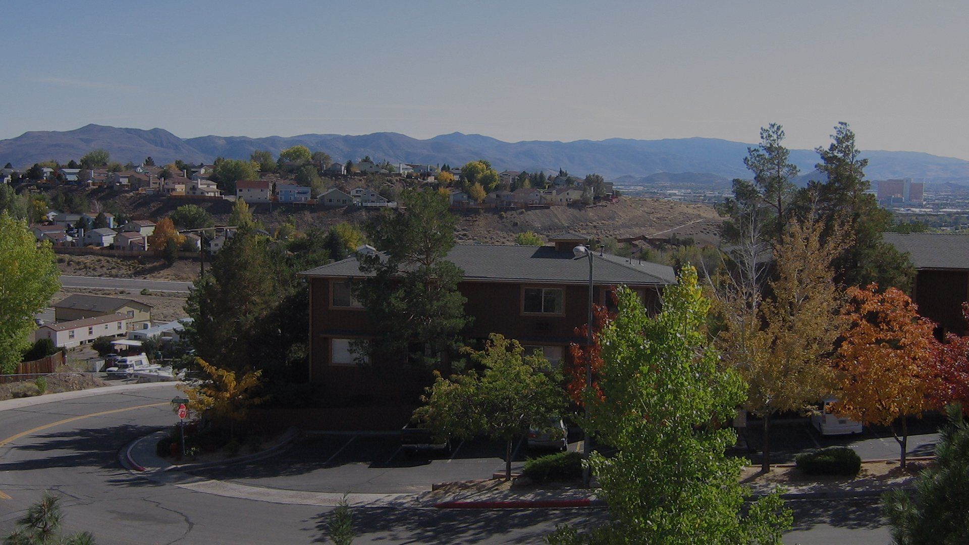 Street-level view of a hillside residential community with a brown building, trees, and distant mountains at Reno Vista Apartments.