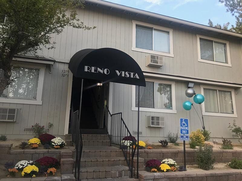 Exterior apartment building entrance with a curved black awning reading 'RENO VISTA,' stairs, and a handicap parking sign at Reno Vista Apartments.