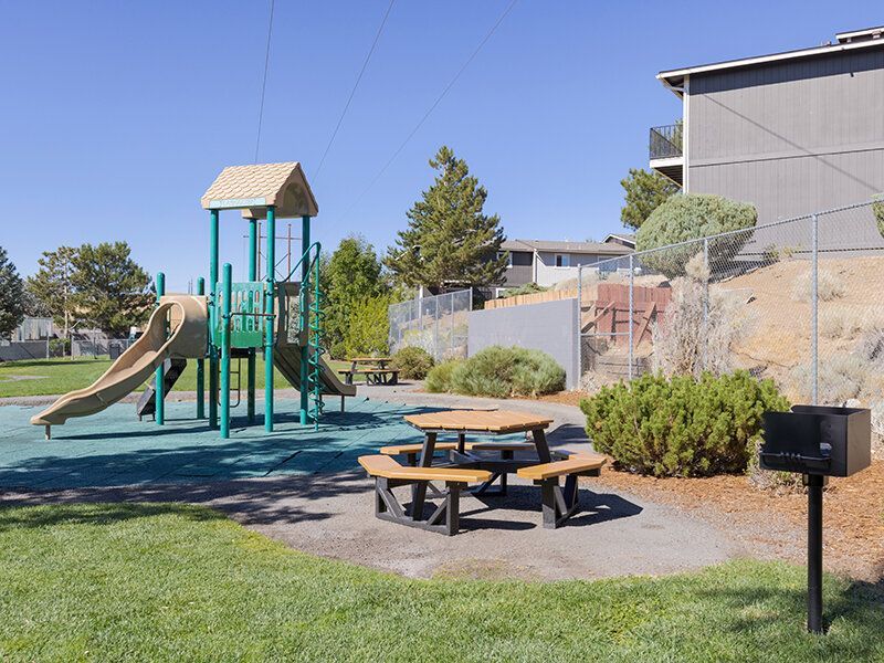 Playground with a slide, climbing structure, and wooden picnic tables with a nearby grill at Reno Vista Apartments.