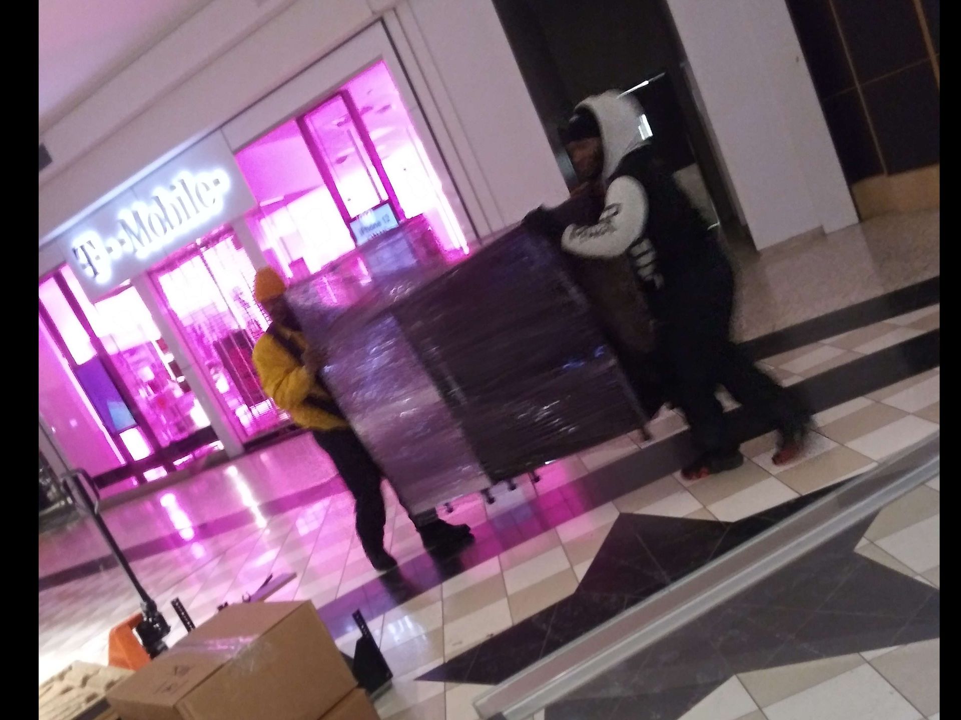 Two people are carrying boxes in a mall in front of a t mobile store