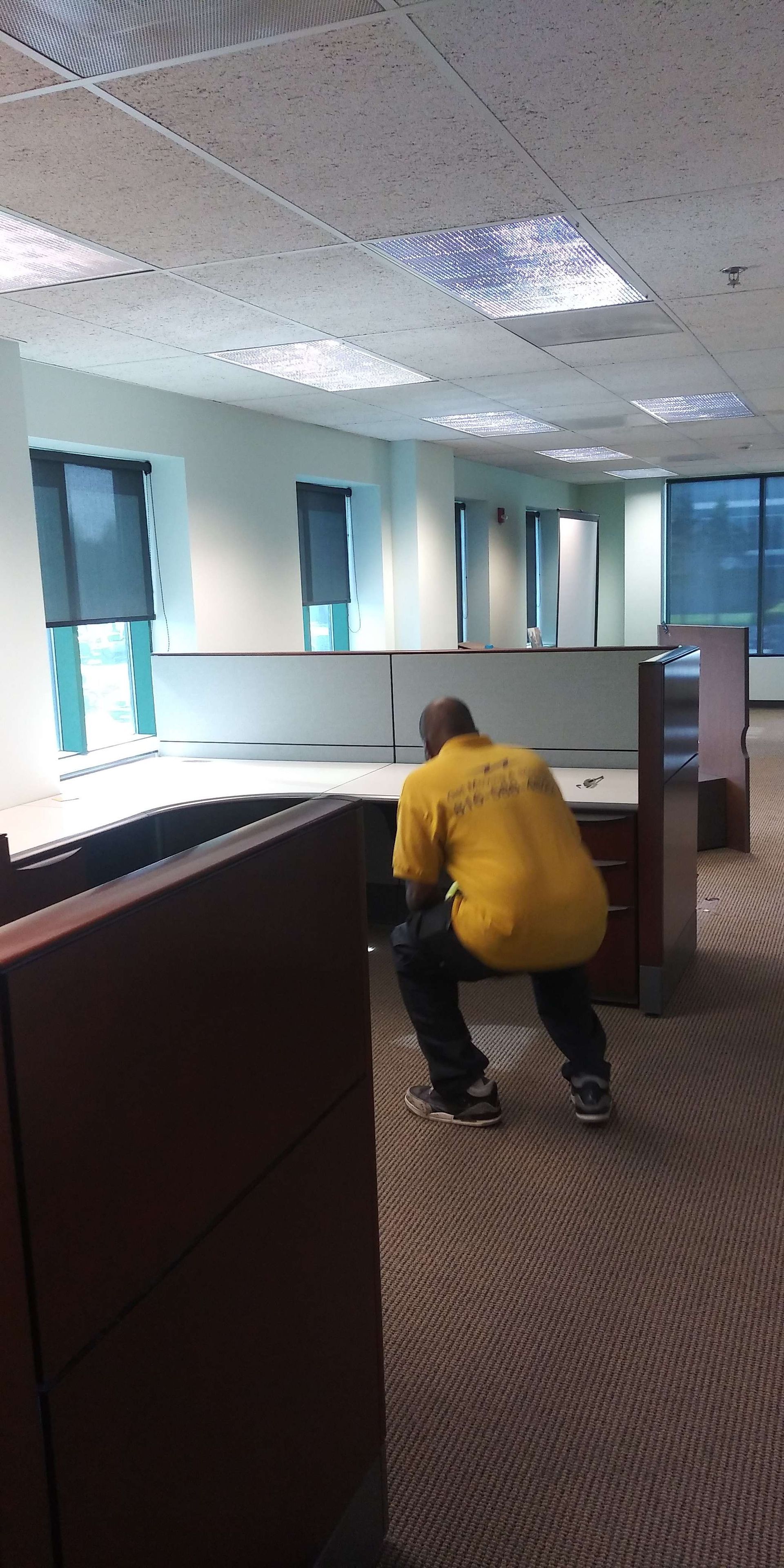 A man in a yellow shirt is kneeling down in an office cubicle.