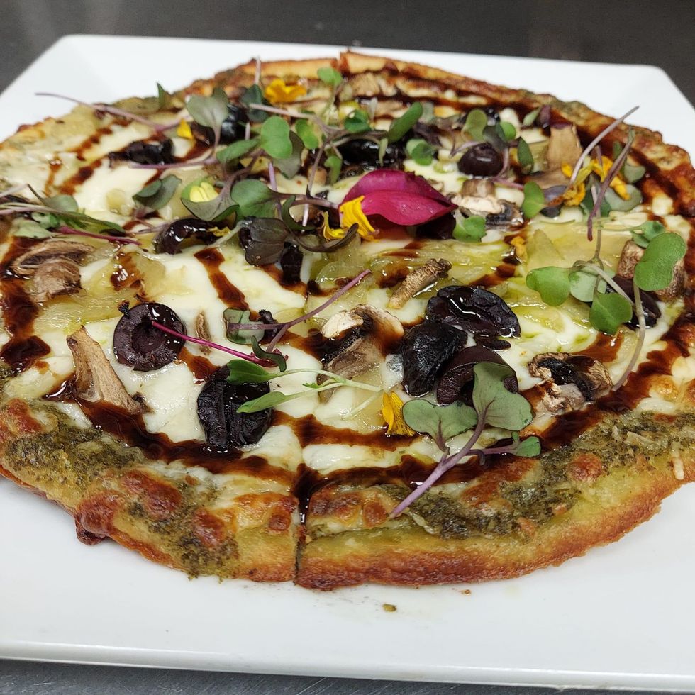 Flatbread pizza on a square white plate, with pesto base, mozzarella, olives, mushrooms, and balsamic glaze.