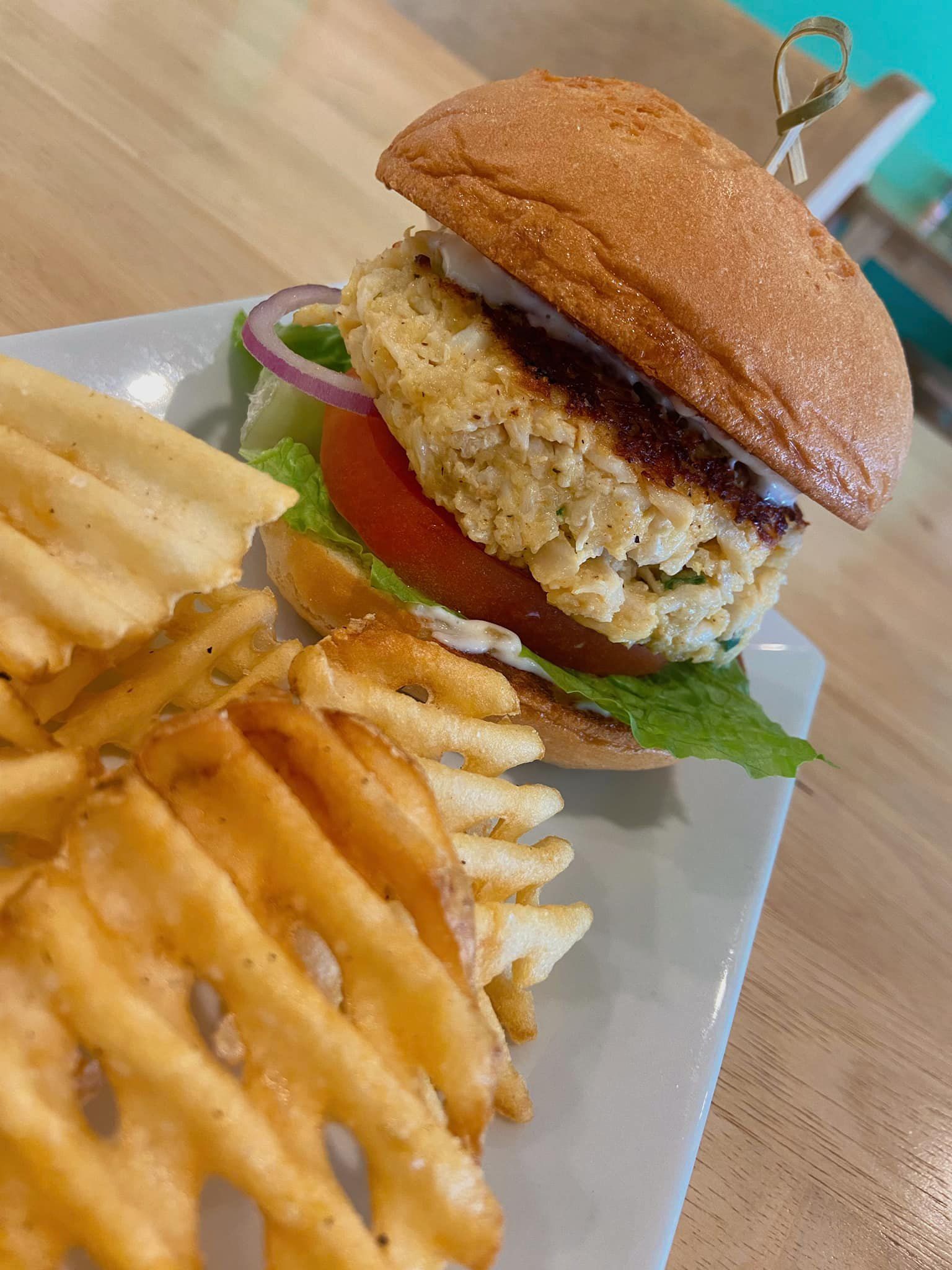 Burger on a bun with lettuce, tomato, and red onion, served with waffle fries on a white plate.