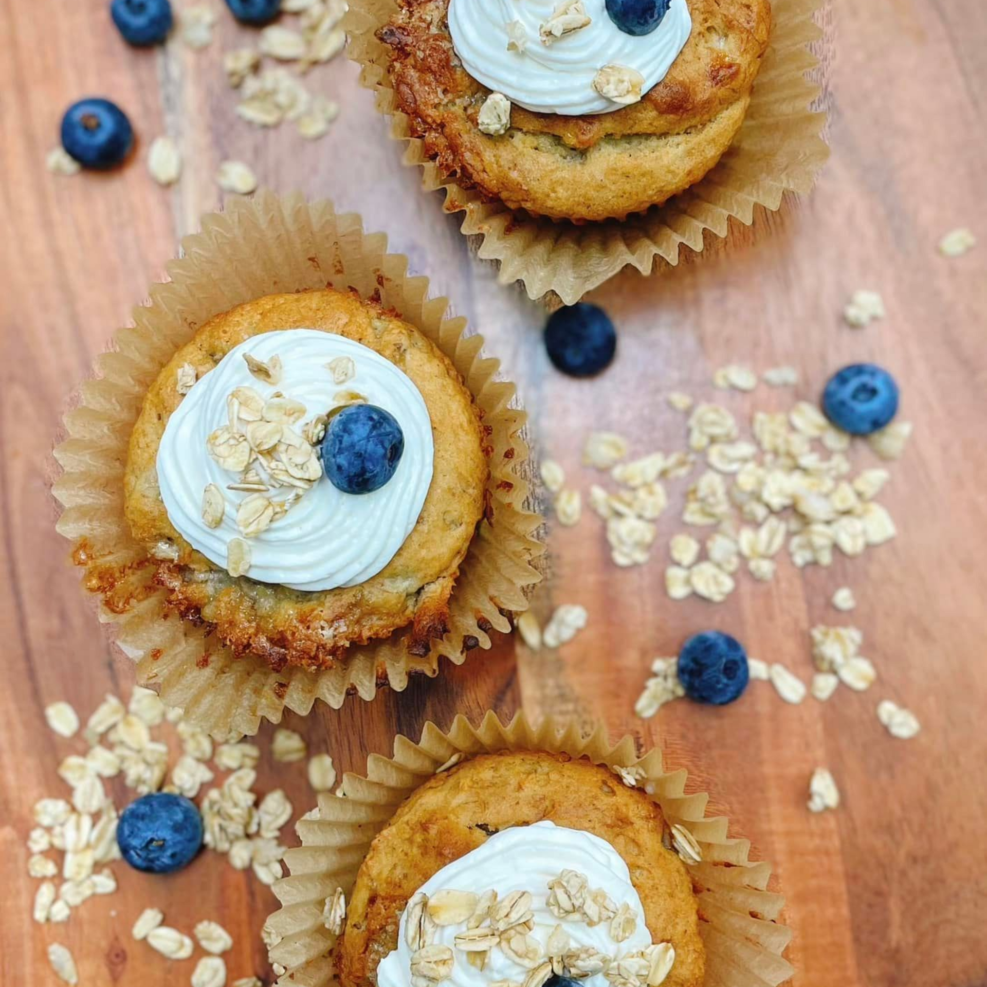 Three muffins in paper liners, topped with white frosting, oats, and blueberries on a wooden surface.