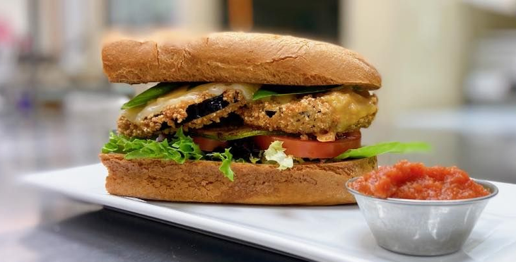 Fried eggplant sandwich with lettuce, tomato, and sauce on a white plate.