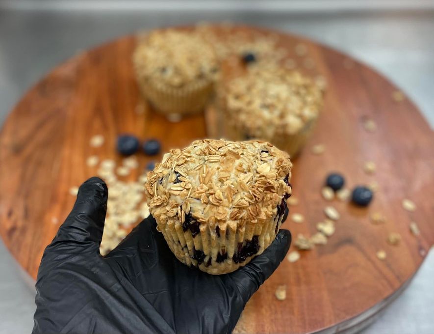 Hand holding a blueberry muffin topped with oats on a wooden board with blueberries and muffins in the background.