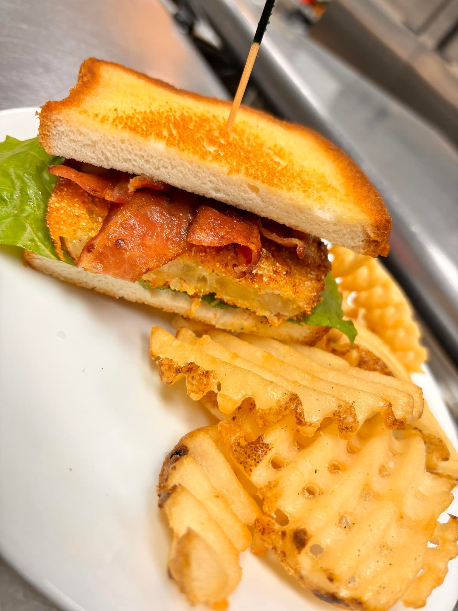 Sandwich with fried tomato, lettuce, and bacon on toasted bread, served with waffle fries.