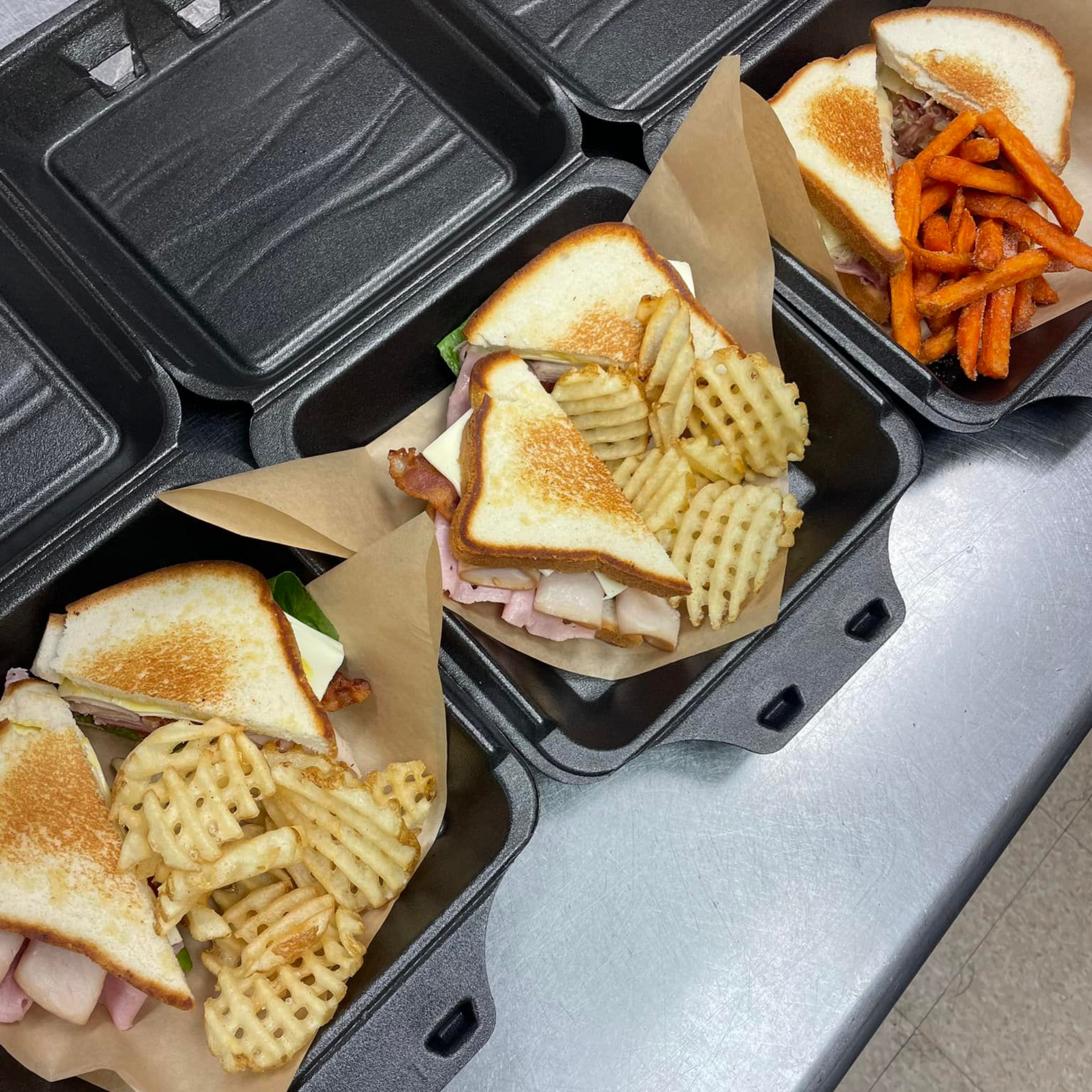 Sandwiches in black takeout containers with waffle fries and sweet potato fries.
