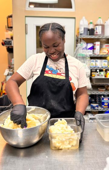Woman wearing black gloves and apron mixes food in a metal bowl, then transfers it to a container.