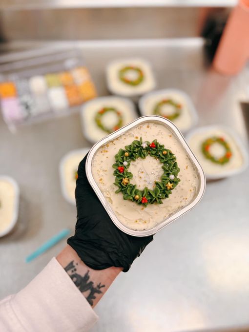 Hand holding a small square cake decorated with a green wreath and gold and red accents. Other cakes in background.