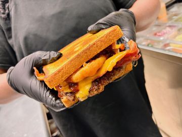 Person holding a large burger on toasted bread with bacon, cheese, and onion rings.